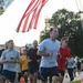 A group of runners participates in an organized run event, likely a military fitness run or memorial run based on the U.S. Air Force insignia visible on several participants' shirts and shorts. An American flag is prominently displayed overhead, suspended from what appears to be a fire truck ladder. Orange traffic cones mark the course route on a paved road.