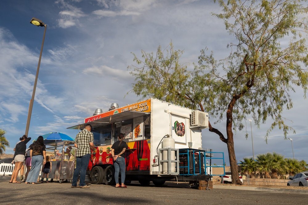 A colorful food trailer is parked in an open lot, likely in a desert Southwest location based on the vegetation and landscape. Several customers are gathered at the serving window, with signage reading 'COME MAKE YOUR [taste buds] TINGLE!' visible on the trailer. The scene appears to be taken during late afternoon with warm sunlight and a partly cloudy sky.