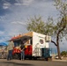 A colorful food trailer is parked in an open lot, likely in a desert Southwest location based on the vegetation and landscape. Several customers are gathered at the serving window, with signage reading 'COME MAKE YOUR [taste buds] TINGLE!' visible on the trailer. The scene appears to be taken during late afternoon with warm sunlight and a partly cloudy sky.