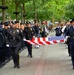 A formal law enforcement ceremonial procession is taking place along a tree-lined walkway, with officers in dark dress uniforms carrying an unfolded American flag. A bagpipe band in traditional Scottish tartan kilts follows behind the flag bearers. Officers line both sides of the path in a formation of honor, with some saluting or recording the event.