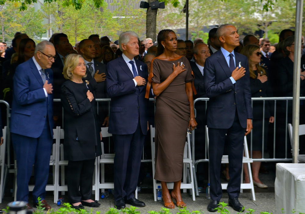 A group of formally dressed individuals stand in a row with their right hands placed over their hearts, suggesting a pledge or national anthem moment. The setting appears to be an outdoor public ceremony with security barriers, a large crowd in the background, and trees visible. The solemn posture and formal attire of the attendees indicate a significant commemorative or official event.