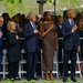 A group of formally dressed individuals stand in a row with their right hands placed over their hearts, suggesting a pledge or national anthem moment. The setting appears to be an outdoor public ceremony with security barriers, a large crowd in the background, and trees visible. The solemn posture and formal attire of the attendees indicate a significant commemorative or official event.