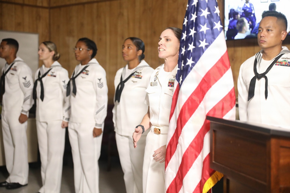 A group of U.S. Navy personnel in white dress uniforms are standing at attention during what appears to be a formal military ceremony. A central figure, likely a senior officer, stands beside an American flag and appears to be speaking or reciting something. The setting is an indoor room with wood-paneled walls and a television monitor visible in the background.