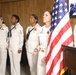 A group of U.S. Navy personnel in white dress uniforms are standing at attention during what appears to be a formal military ceremony. A central figure, likely a senior officer, stands beside an American flag and appears to be speaking or reciting something. The setting is an indoor room with wood-paneled walls and a television monitor visible in the background.
