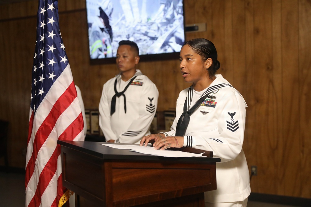 Two U.S. Navy sailors in white dress uniforms are participating in a formal ceremony or presentation. A female sailor with the rank of Petty Officer First Class stands at a wooden podium speaking, while a male sailor stands at attention in the background. An American flag is prominently displayed to the left, and a screen showing what appears to be disaster or rescue imagery is visible in the background.