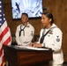 Two U.S. Navy sailors in white dress uniforms are participating in a formal ceremony or presentation. A female sailor with the rank of Petty Officer First Class stands at a wooden podium speaking, while a male sailor stands at attention in the background. An American flag is prominently displayed to the left, and a screen showing what appears to be disaster or rescue imagery is visible in the background.