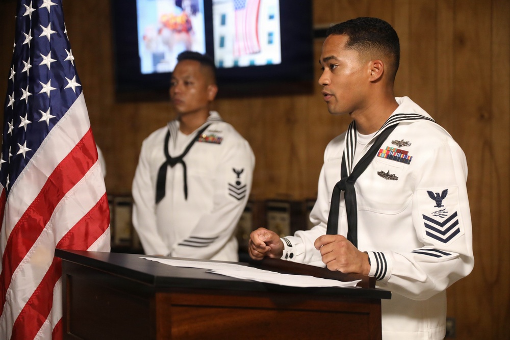 Two U.S. Navy sailors in dress white uniforms are participating in a formal military ceremony or event. The sailor in the foreground is speaking at a wooden podium with documents visible, while a second sailor stands at attention in the background. An American flag is prominently displayed to the left, and a monitor showing patriotic imagery is visible in the background.