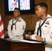 Two U.S. Navy sailors in dress white uniforms are participating in a formal military ceremony or event. The sailor in the foreground is speaking at a wooden podium with documents visible, while a second sailor stands at attention in the background. An American flag is prominently displayed to the left, and a monitor showing patriotic imagery is visible in the background.