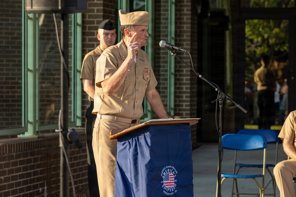 A U.S. Navy officer in khaki service uniform is delivering a speech at a podium during an outdoor ceremony at Norfolk Naval Shipyard in Portsmouth, Virginia. The podium is draped with a blue cover bearing the Norfolk Naval Shipyard logo and the text 'America's Shipyard, Portsmouth, VA.' A second uniformed service member stands at attention in the background.