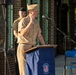 A U.S. Navy officer in khaki service uniform is delivering a speech at a podium during an outdoor ceremony at Norfolk Naval Shipyard in Portsmouth, Virginia. The podium is draped with a blue cover bearing the Norfolk Naval Shipyard logo and the text 'America's Shipyard, Portsmouth, VA.' A second uniformed service member stands at attention in the background.