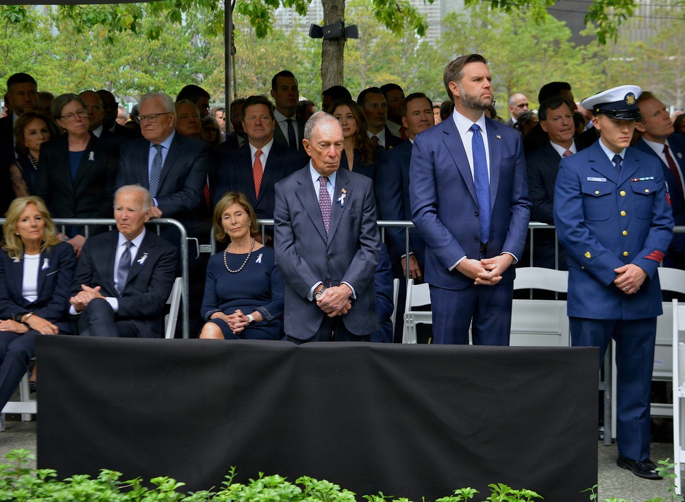 A formal outdoor memorial or commemorative ceremony is taking place, with a large group of attendees dressed in dark, formal attire. The setting appears to be an official public event, with rows of seated and standing attendees, a black draped barrier in the foreground, and green trees in the background. A uniformed military or coast guard officer is visible on the right side of the image.