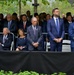 A formal outdoor memorial or commemorative ceremony is taking place, with a large group of attendees dressed in dark, formal attire. The setting appears to be an official public event, with rows of seated and standing attendees, a black draped barrier in the foreground, and green trees in the background. A uniformed military or coast guard officer is visible on the right side of the image.