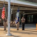 A military color guard ceremony is taking place outdoors in front of a brick building entrance. Personnel in tan/khaki uniforms are presenting the American flag and what appears to be a U.S. Navy flag, while additional service members are seated in the background. A wooden podium with a bell is visible on the right side of the formation.