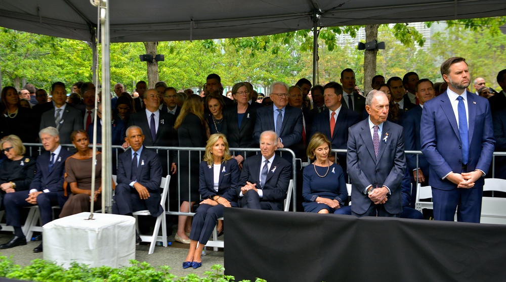 A formal outdoor memorial or commemorative ceremony is taking place under a large tent canopy, with a large crowd of attendees dressed in formal dark attire. The gathering appears solemn in nature, with attendees seated and standing in organized rows. White ribbons or pins are visible on several attendees, suggesting a memorial event.