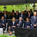A formal outdoor memorial or commemorative ceremony is taking place under a large tent canopy, with a large crowd of attendees dressed in formal dark attire. The gathering appears solemn in nature, with attendees seated and standing in organized rows. White ribbons or pins are visible on several attendees, suggesting a memorial event.