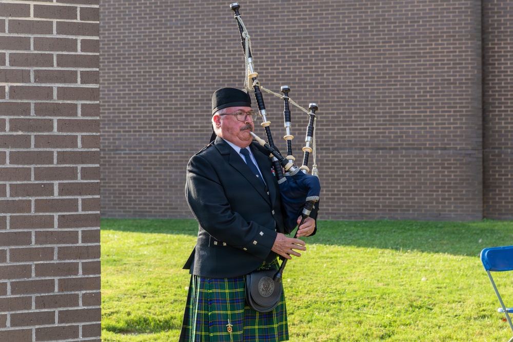A middle-aged man dressed in traditional Scottish Highland attire is playing bagpipes outdoors near a brick building. He is wearing a dark jacket, a green and blue tartan kilt, and a black cap. The setting appears to be a formal or ceremonial outdoor event.
