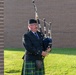 A middle-aged man dressed in traditional Scottish Highland attire is playing bagpipes outdoors near a brick building. He is wearing a dark jacket, a green and blue tartan kilt, and a black cap. The setting appears to be a formal or ceremonial outdoor event.
