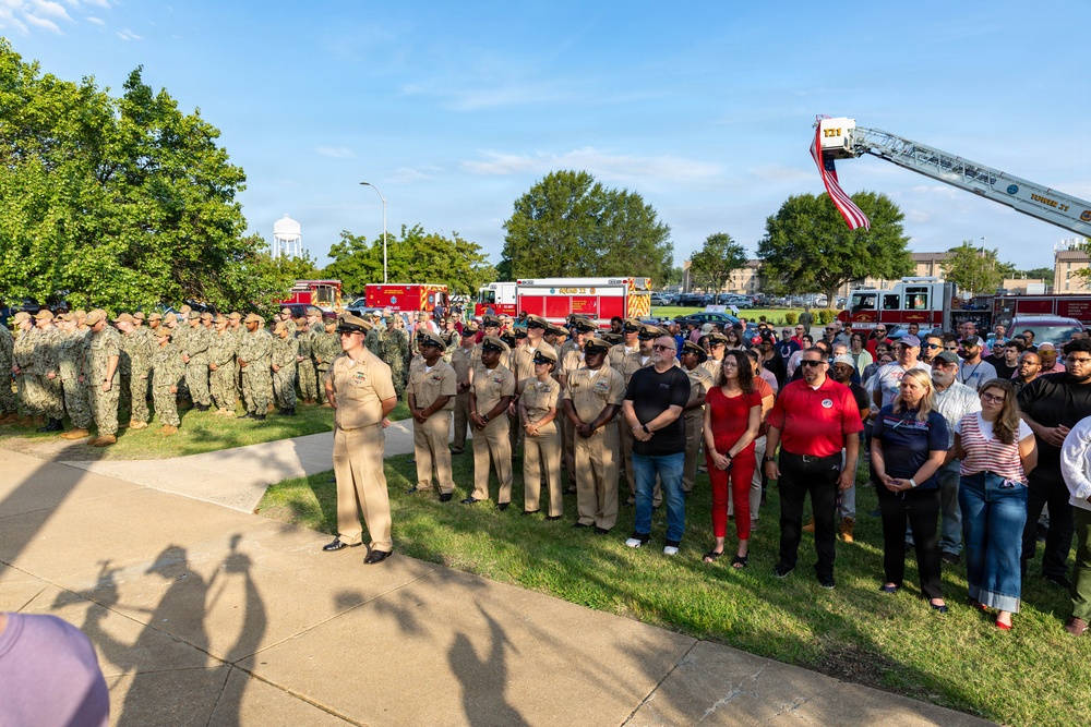 A large outdoor military ceremony or memorial gathering featuring U.S. Navy personnel in both dress khaki and camouflage uniforms, alongside civilian attendees. A fire truck aerial ladder is extended in the background displaying an American flag, suggesting a patriotic or commemorative event. The gathering appears to take place on a military installation, likely a naval base.