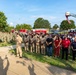 A large outdoor military ceremony or memorial gathering featuring U.S. Navy personnel in both dress khaki and camouflage uniforms, alongside civilian attendees. A fire truck aerial ladder is extended in the background displaying an American flag, suggesting a patriotic or commemorative event. The gathering appears to take place on a military installation, likely a naval base.