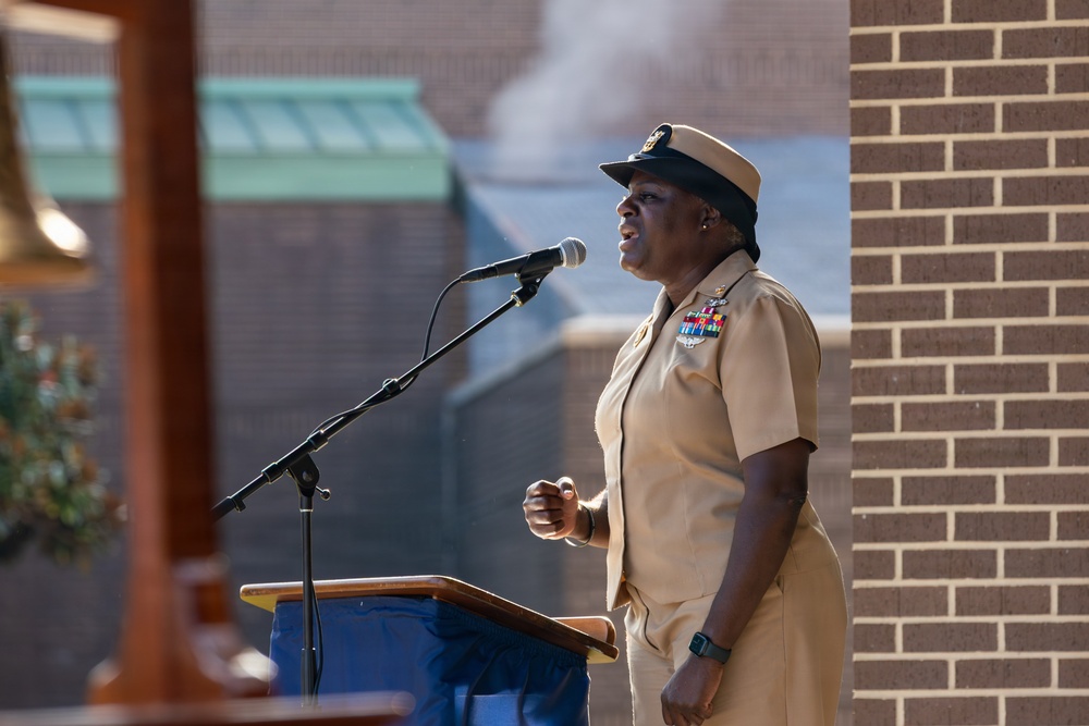 A U.S. Navy senior enlisted officer in khaki service uniform is speaking at a podium with a microphone at what appears to be an outdoor military ceremony. She is wearing multiple service ribbons and a command insignia on her uniform, along with the distinctive Navy combination cover (cap). The setting appears to be a military installation, with a brick building visible in the background.