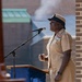 A U.S. Navy senior enlisted officer in khaki service uniform is speaking at a podium with a microphone at what appears to be an outdoor military ceremony. She is wearing multiple service ribbons and a command insignia on her uniform, along with the distinctive Navy combination cover (cap). The setting appears to be a military installation, with a brick building visible in the background.