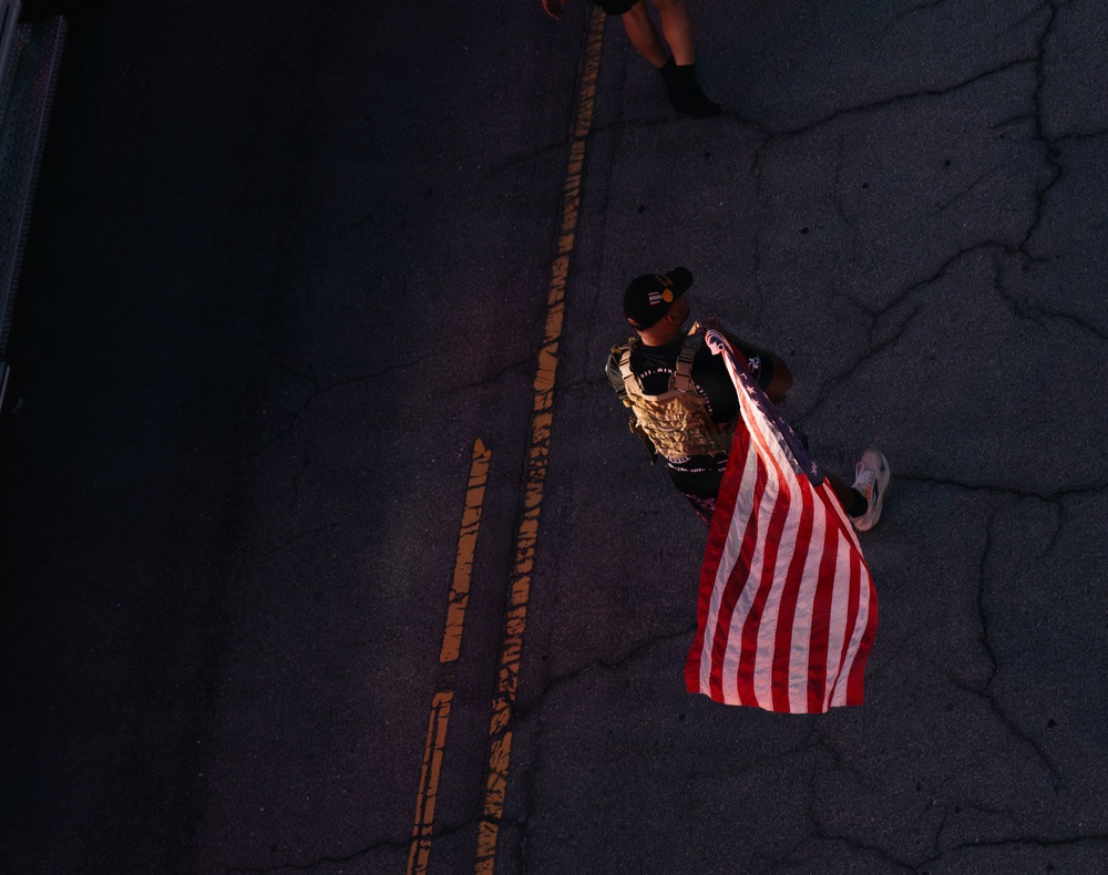 An aerial/overhead photograph taken from above shows a person walking on a road carrying an American flag. The individual is wearing tactical/military-style gear including a vest and a cap, and is walking along a road with yellow center line markings. A second person is partially visible at the top of the frame, visible only from the lower legs down.