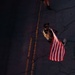 An aerial/overhead photograph taken from above shows a person walking on a road carrying an American flag. The individual is wearing tactical/military-style gear including a vest and a cap, and is walking along a road with yellow center line markings. A second person is partially visible at the top of the frame, visible only from the lower legs down.