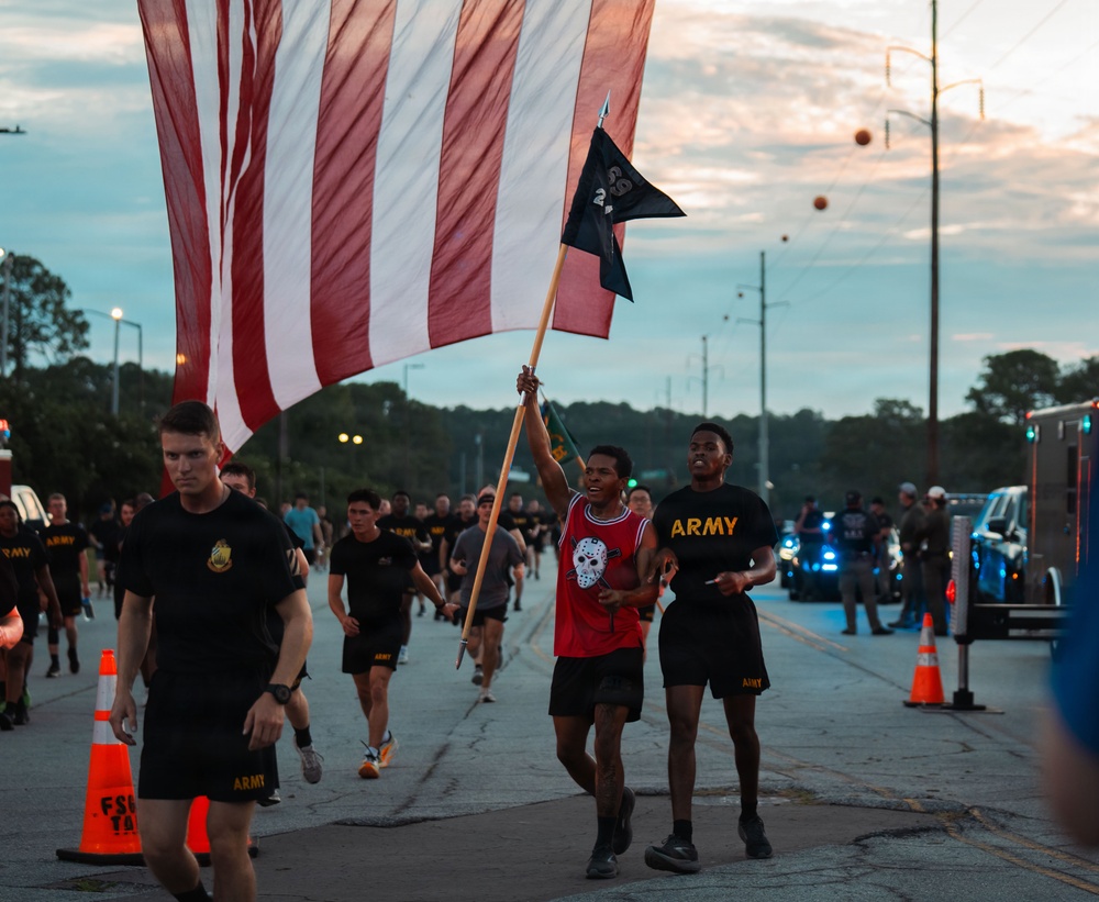A group of U.S. Army soldiers and participants are running along a road during what appears to be an early morning or dusk formation run or memorial event. A central figure in a red jersey is carrying a large American flag alongside a smaller black pennant flag. The scene is organized with traffic cones lining the route and a police/emergency vehicle with blue lights visible in the background.