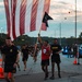 A group of U.S. Army soldiers and participants are running along a road during what appears to be an early morning or dusk formation run or memorial event. A central figure in a red jersey is carrying a large American flag alongside a smaller black pennant flag. The scene is organized with traffic cones lining the route and a police/emergency vehicle with blue lights visible in the background.