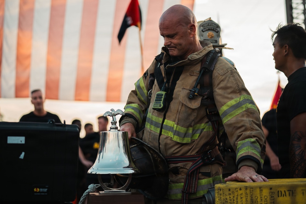 A firefighter in full turnout gear stands solemnly over a ceremonial bell, likely during a memorial or tribute event. An American flag is visible in the background along with other attendees dressed in black. The scene has the hallmarks of a fallen firefighter memorial ceremony.