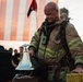 A firefighter in full turnout gear stands solemnly over a ceremonial bell, likely during a memorial or tribute event. An American flag is visible in the background along with other attendees dressed in black. The scene has the hallmarks of a fallen firefighter memorial ceremony.