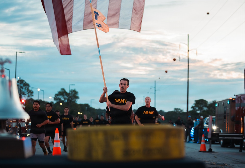 A group of U.S. Army soldiers in black PT (physical training) uniforms are running in formation along a road at dusk or dawn. The lead soldier is carrying what appears to be an American flag and a unit guidon on a wooden pole. The scene appears to be a military formation run or physical training event, with traffic cones and emergency vehicles visible along the route.