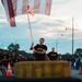 A group of U.S. Army soldiers in black PT (physical training) uniforms are running in formation along a road at dusk or dawn. The lead soldier is carrying what appears to be an American flag and a unit guidon on a wooden pole. The scene appears to be a military formation run or physical training event, with traffic cones and emergency vehicles visible along the route.