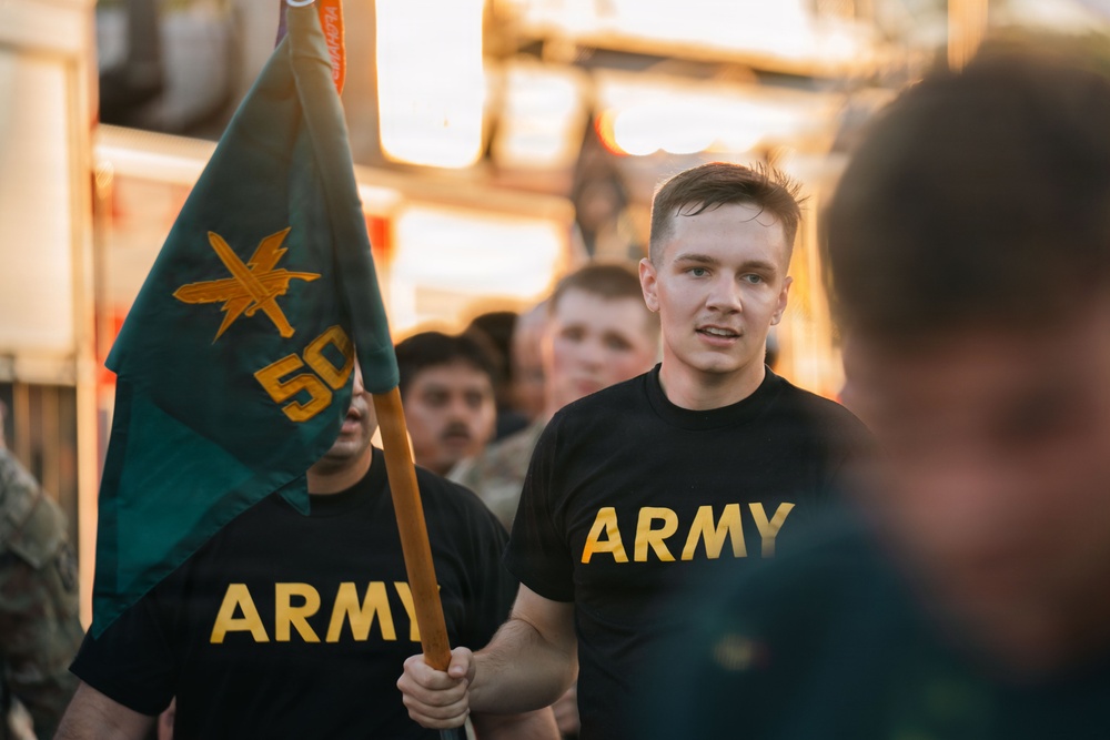A group of U.S. Army soldiers or trainees are marching or running in formation, wearing black PT (physical training) shirts with gold 'ARMY' lettering. One individual in the foreground is carrying a teal/green guidon flag bearing a gold insignia and the number '50'. The image appears to capture a military formation run or ceremony event.