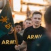 A group of U.S. Army soldiers or trainees are marching or running in formation, wearing black PT (physical training) shirts with gold 'ARMY' lettering. One individual in the foreground is carrying a teal/green guidon flag bearing a gold insignia and the number '50'. The image appears to capture a military formation run or ceremony event.