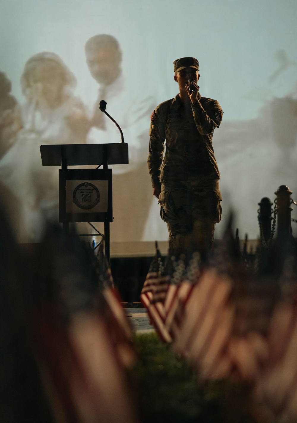 A soldier in military camouflage uniform and patrol cap stands on a stage holding a handheld microphone, appearing to sing or speak at a formal military ceremony. A podium with an emblem is visible to the left, and rows of small American flags are arranged in the foreground. A large projection screen in the background casts shadows of the performer.