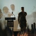 A soldier in military camouflage uniform and patrol cap stands on a stage holding a handheld microphone, appearing to sing or speak at a formal military ceremony. A podium with an emblem is visible to the left, and rows of small American flags are arranged in the foreground. A large projection screen in the background casts shadows of the performer.