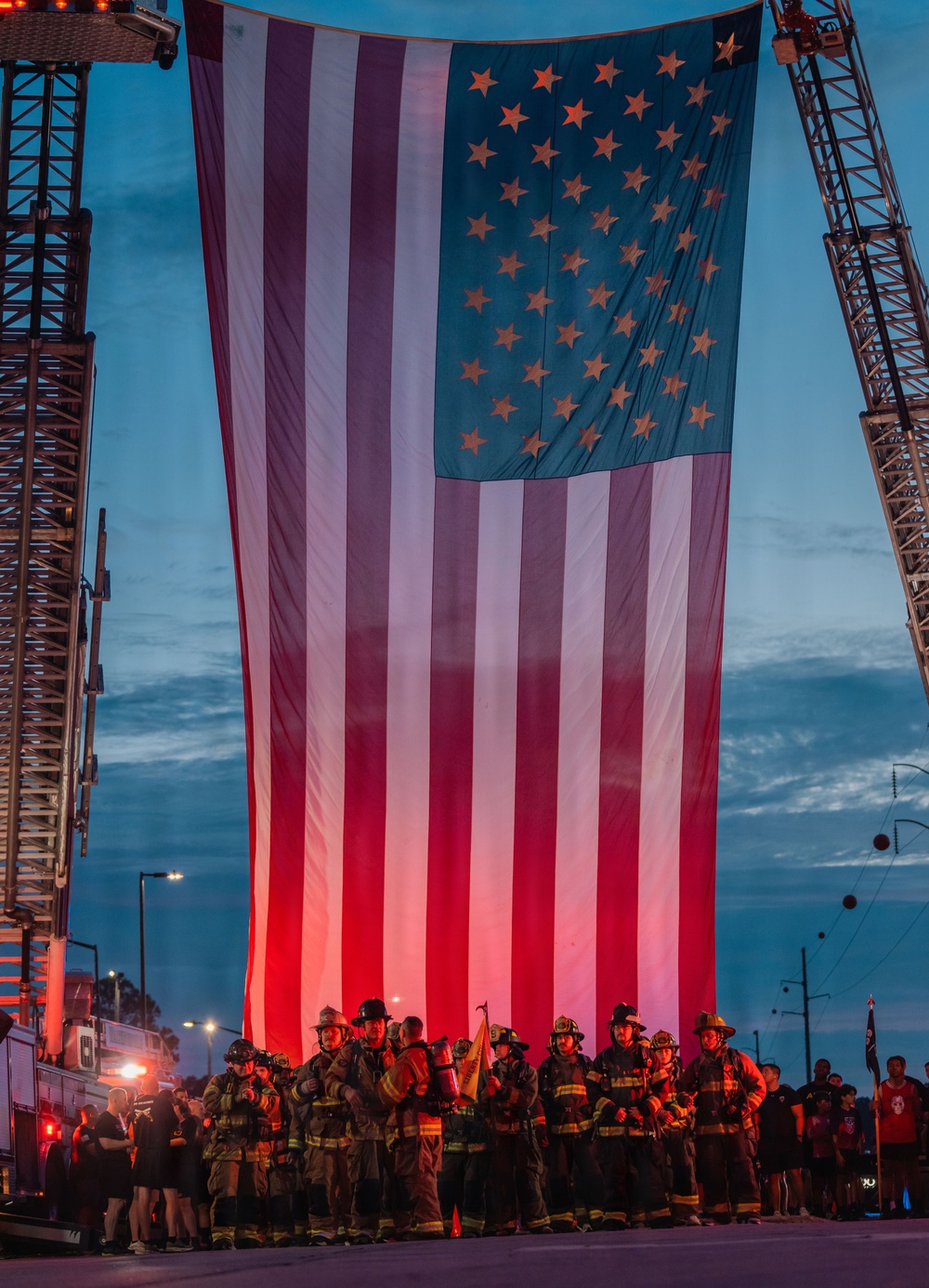 A large American flag is suspended between two fire truck aerial ladders at dusk or dawn, illuminated from below with red light. A group of firefighters in full turnout gear stand assembled beneath the flag in formation. The scene has the appearance of a memorial or ceremonial event, likely a 9/11 remembrance or fallen firefighter tribute.