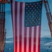 A large American flag is suspended between two fire truck aerial ladders at dusk or dawn, illuminated from below with red light. A group of firefighters in full turnout gear stand assembled beneath the flag in formation. The scene has the appearance of a memorial or ceremonial event, likely a 9/11 remembrance or fallen firefighter tribute.