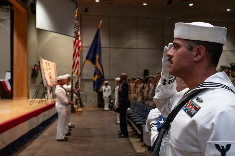 A formal U.S. Navy military ceremony is taking place in an auditorium or conference hall. A sailor in dress white uniform is prominently saluting in the foreground, wearing multiple service ribbons and a white cover (hat). In the background, a color guard holds the American flag and a blue military branch flag near a stage, with rows of personnel in various uniforms standing at attention.