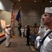 A formal U.S. Navy military ceremony is taking place in an auditorium or conference hall. A sailor in dress white uniform is prominently saluting in the foreground, wearing multiple service ribbons and a white cover (hat). In the background, a color guard holds the American flag and a blue military branch flag near a stage, with rows of personnel in various uniforms standing at attention.