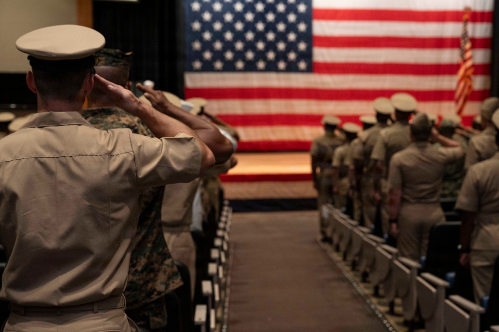 Military personnel in various uniforms are standing at attention and saluting a large American flag displayed on a stage in what appears to be an auditorium or ceremony hall. The scene depicts a formal military ceremony with service members arranged in rows facing the flag. At least two distinct uniform types are visible, suggesting a joint-service or mixed-rank event.