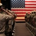 Military personnel in various uniforms are standing at attention and saluting a large American flag displayed on a stage in what appears to be an auditorium or ceremony hall. The scene depicts a formal military ceremony with service members arranged in rows facing the flag. At least two distinct uniform types are visible, suggesting a joint-service or mixed-rank event.