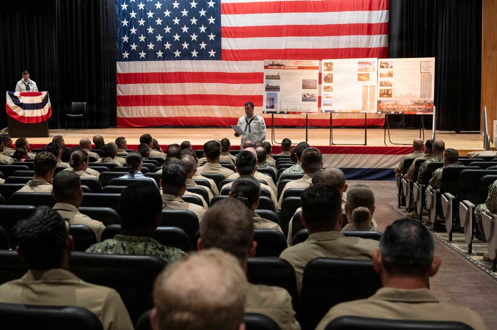 A military remembrance ceremony is taking place in an auditorium or theater, with uniformed service members seated in rows facing a stage. A large American flag serves as a backdrop on the stage, where a sailor in dress whites stands speaking, flanked by informational display boards. The display boards appear to reference September 11, 2001 remembrance based on visible text and imagery.