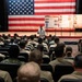A military remembrance ceremony is taking place in an auditorium or theater, with uniformed service members seated in rows facing a stage. A large American flag serves as a backdrop on the stage, where a sailor in dress whites stands speaking, flanked by informational display boards. The display boards appear to reference September 11, 2001 remembrance based on visible text and imagery.