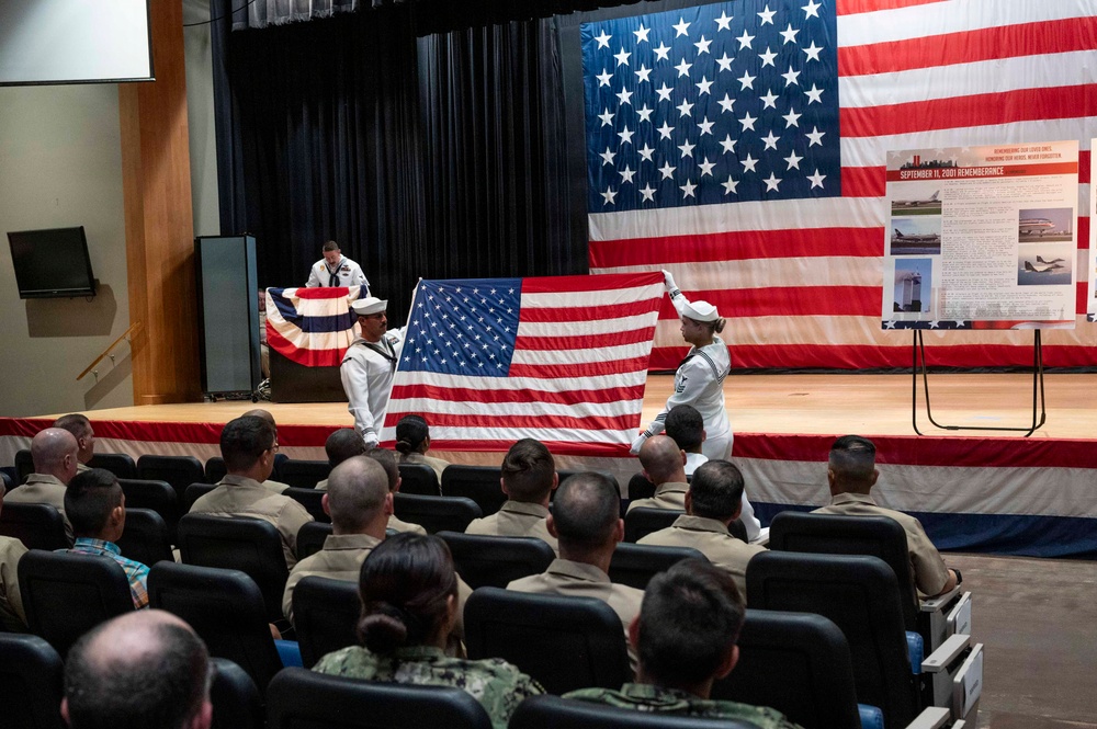 A military ceremony, specifically a September 11, 2001 Remembrance event, is taking place in an auditorium or theater. Two Navy sailors in dress whites are unfolding or presenting an American flag on stage in front of a large backdrop American flag. An audience of military personnel in tan/khaki uniforms is seated watching the ceremony.