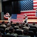 A military ceremony, specifically a September 11, 2001 Remembrance event, is taking place in an auditorium or theater. Two Navy sailors in dress whites are unfolding or presenting an American flag on stage in front of a large backdrop American flag. An audience of military personnel in tan/khaki uniforms is seated watching the ceremony.