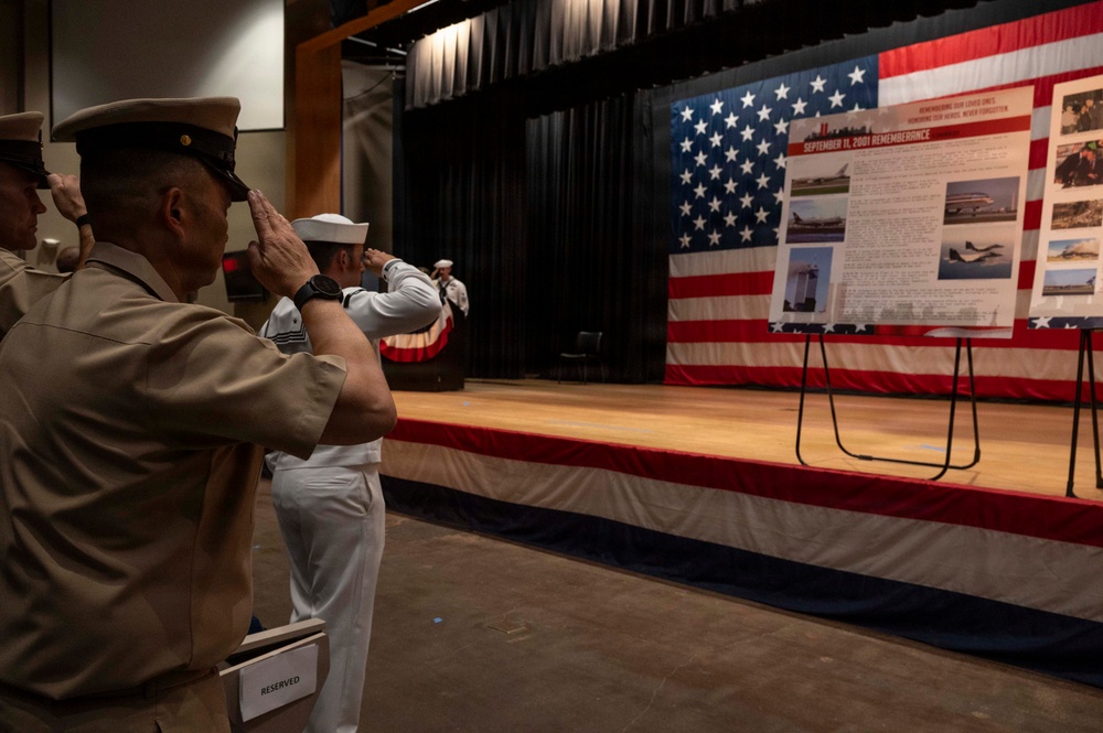 Military personnel in various uniforms are saluting during a September 11, 2001 Remembrance ceremony. The event takes place in an auditorium-style venue decorated with a large American flag backdrop on a stage. A display board titled 'September 11, 2001 Rememberance' with photographs and text is prominently featured on the stage.