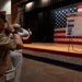 Military personnel in various uniforms are saluting during a September 11, 2001 Remembrance ceremony. The event takes place in an auditorium-style venue decorated with a large American flag backdrop on a stage. A display board titled 'September 11, 2001 Rememberance' with photographs and text is prominently featured on the stage.