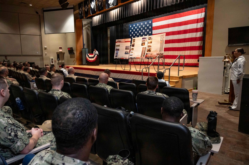 A military briefing or ceremony is taking place in an auditorium, with a large audience of uniformed Navy personnel seated in rows facing a stage. The stage is decorated with a large American flag as a backdrop, along with display boards containing photographs and text. A speaker stands at a podium on stage while a Navy petty officer in dress whites stands at attention near a ship's bell on the right side.